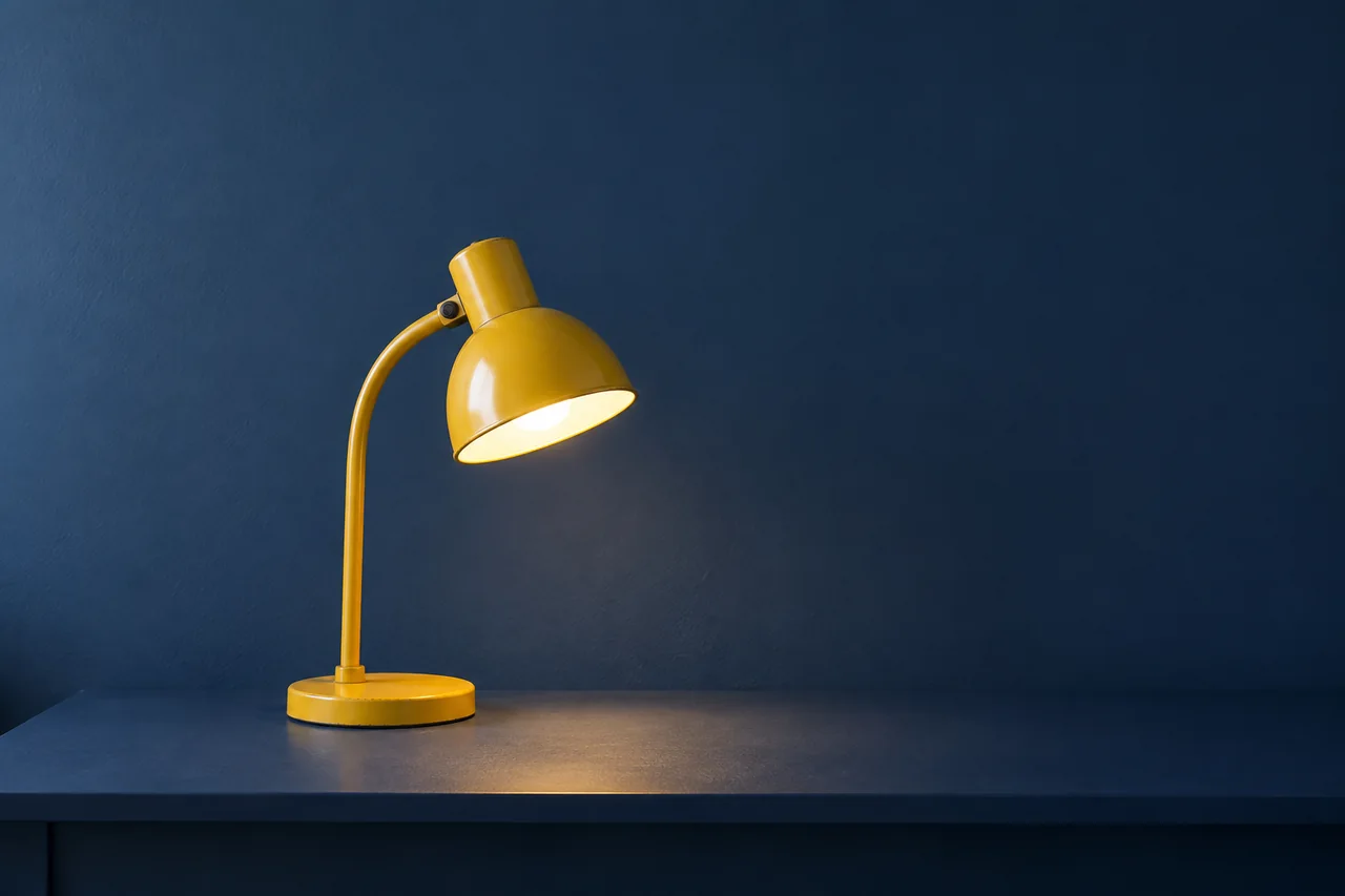 Yellow metal desk lamp with a round base on a navy desk and clear wall space above.